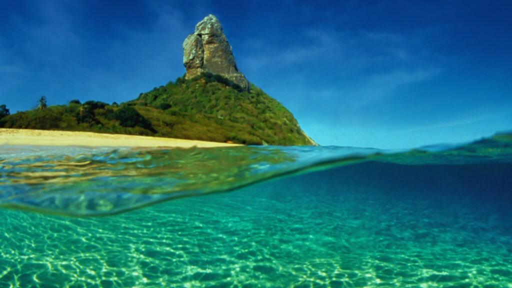 Praia da Conceição em Fernando de Noronha, com Morro do Pico ao fundo e mar de águas cristalinas.