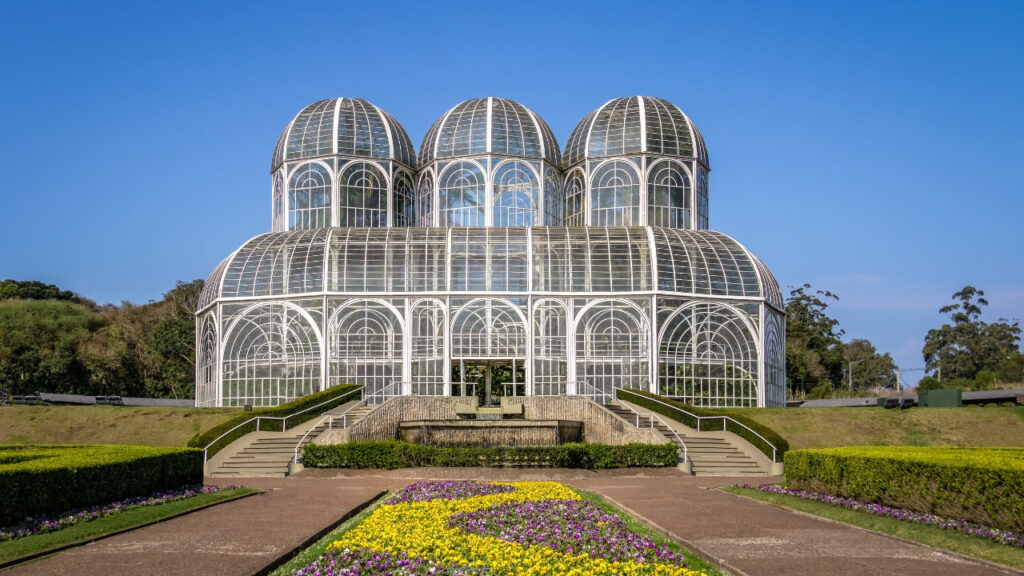 Foto do Jardim Botânico de Curitiba com a estufa de vidro ao centro e jardins floridos ao redor.