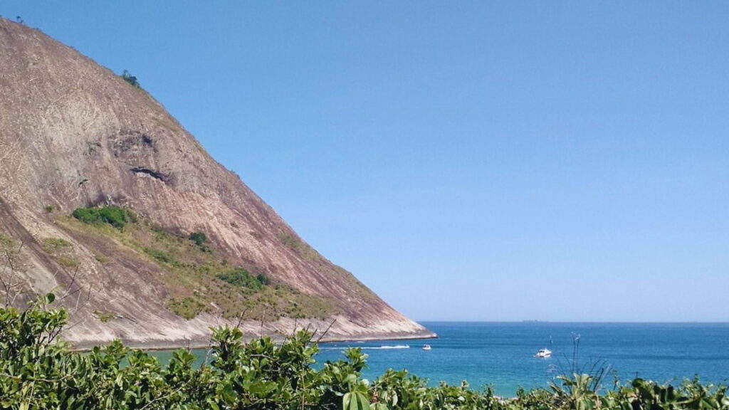 Vista da Praia de Itacoatiara em Niterói, com mar azul, faixa de areia clara, Costão de Itacoatiara ao fundo e pessoas na orla.