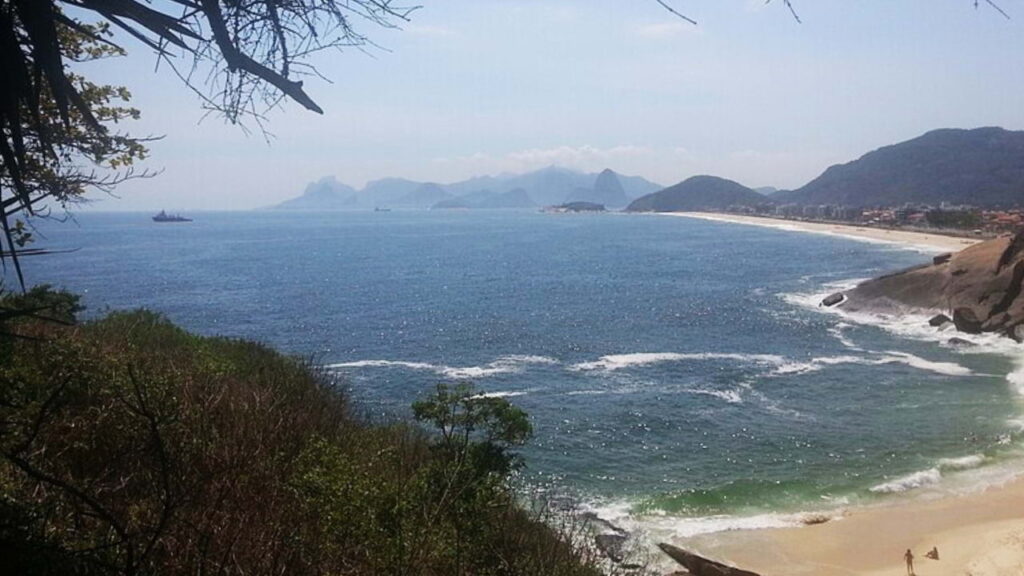 Praia do Sossego em Niterói, com faixa de areia pequena, mar transparente, vegetação nativa ao redor e acesso por trilha entre as pedras.