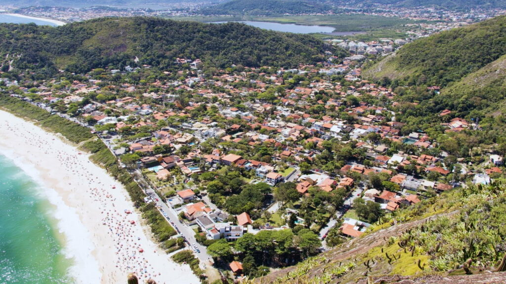 Vista aérea da Praia de Itacoatiara em Niterói, com areia clara, mar azul e o bairro residencial ao fundo cercado por áreas verdes.
