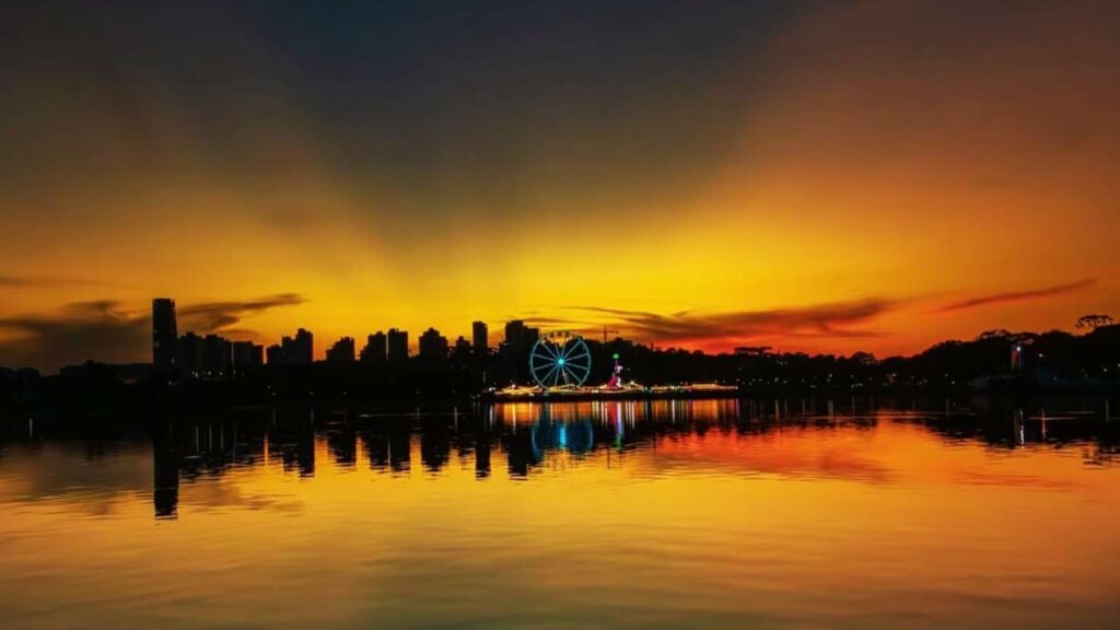 Roda-gigante iluminada no Parque Barigui durante o pôr do sol, refletida no lago, com a silhueta de prédios ao fundo em Curitiba.