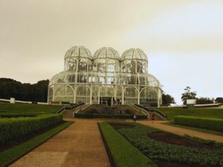 Imagem do Jardim Botânico de Curitiba em dia de chuva, com destaque para a estufa de vidro e jardins.