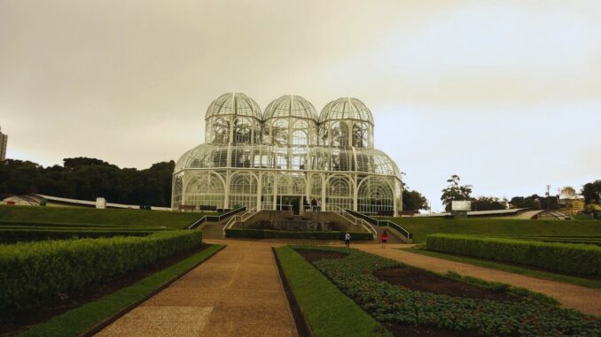 Imagem do Jardim Botânico de Curitiba em dia de chuva, com destaque para a estufa de vidro e jardins.