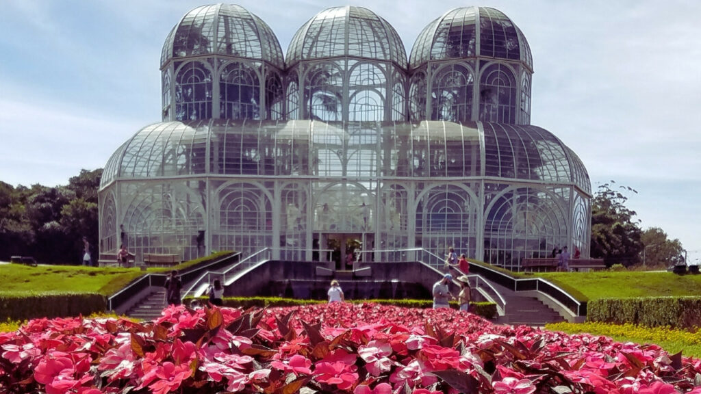 Jardim Botânico de Curitiba na primavera, com estufa de vidro e canteiro florido em primeiro plano.