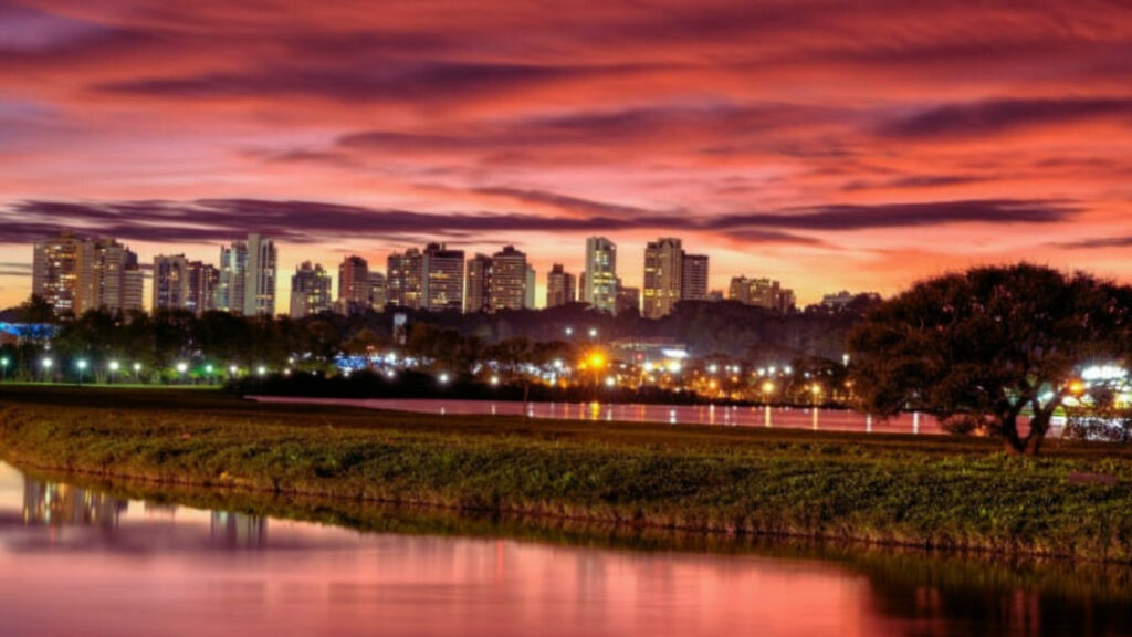 Anoitecer no Parque Barigui com céu avermelhado, lago refletindo as luzes da pista e prédios do Campina do Siqueira ao fundo.