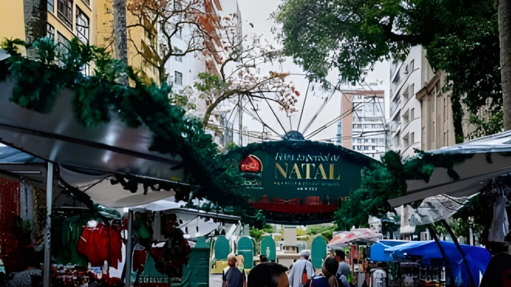 Feira na Praça Santos Andrade, em Curitiba, com barracas iluminadas, artesanato local e visitantes passeando.