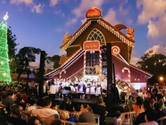 Público reunido no Bosque Alemão, em Curitiba, assistindo a espetáculo natalino ao ar livre com casa temática iluminada e grande árvore de Natal verde.