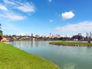 Vista panorâmica do lago do Parque Barigui em Curitiba, com área verde e skyline ao fundo.