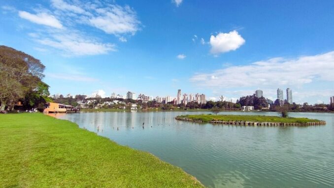 Vista panorâmica do lago do Parque Barigui em Curitiba, com área verde e skyline ao fundo.