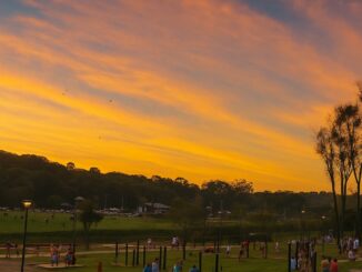 Pôr do sol no Parque Barigui em Curitiba, com o céu alaranjado refletindo no parque e pessoas caminhando e aproveitando o fim de tarde.