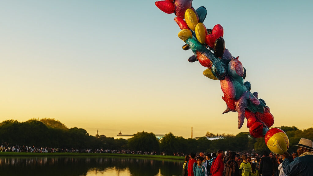 Vendedor de balões no Parque Barigui durante o fim da tarde, horário de maior movimento, com lago e visitantes reunidos ao redor.