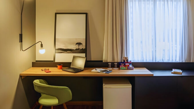 Quarto de hotel em Curitiba com luminária acesa, notebook sobre a mesa e ambiente iluminado, ilustrando o uso da voltagem de 110 volts na cidade.