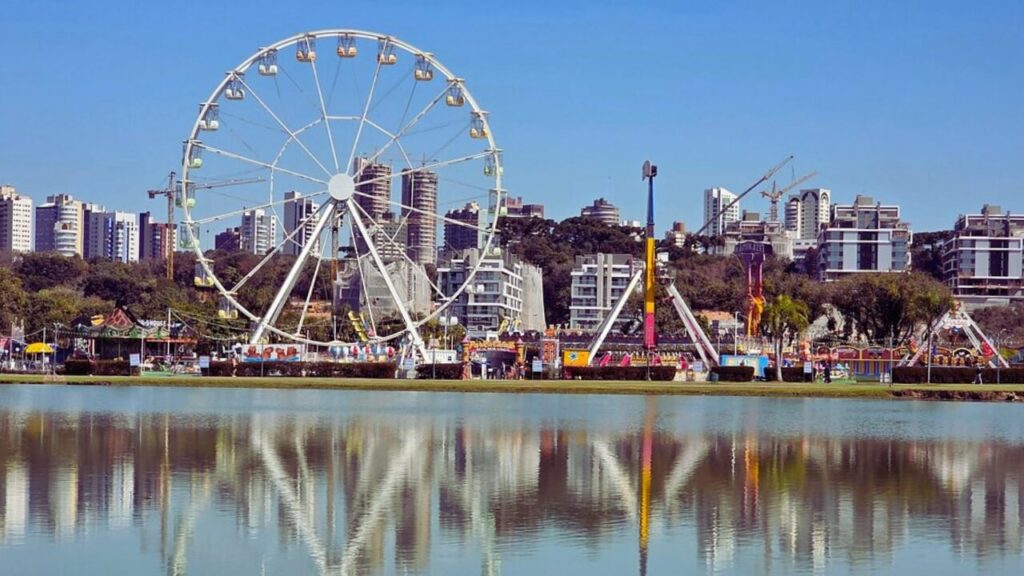 Roda-gigante instalada no Parque Barigui, refletida no lago, com prédios de Curitiba ao fundo.